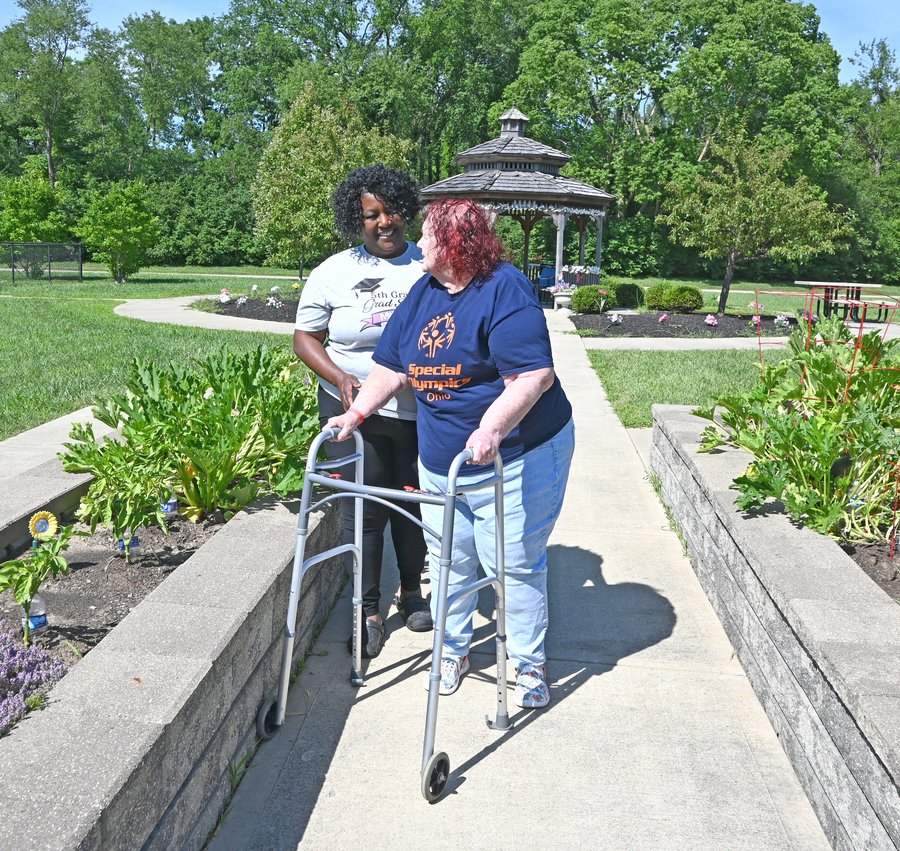 Two people walking together on a concrete path through a sunny garden park in Ohio, with one person using a walker for mobility support