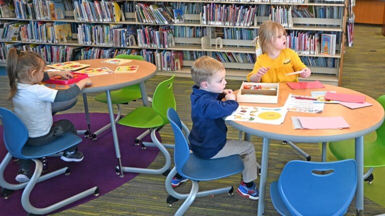 Three young children engaged in creative activities at a round table in a library, with bookshelves in the background