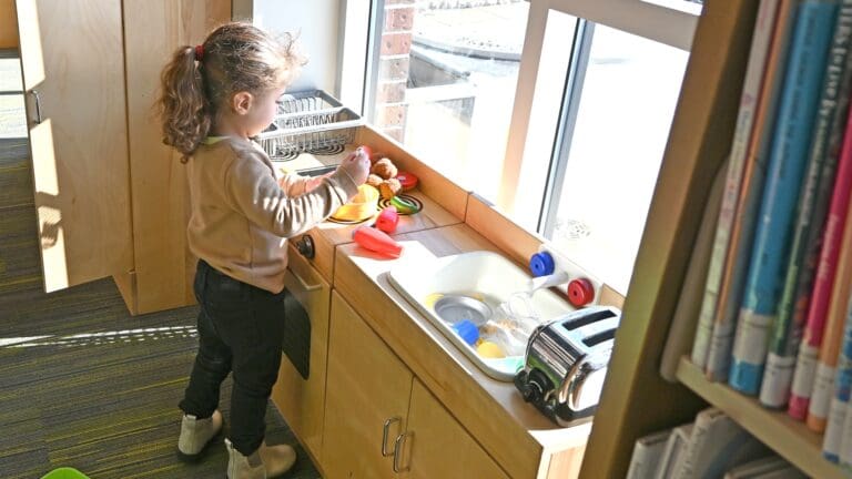 Young child with curly hair playing with toys at a sunny window counter in a classroom or library setting