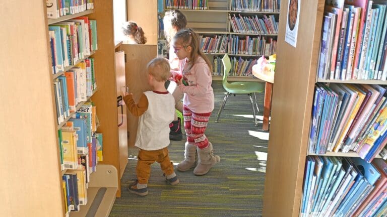Young children exploring books on shelves in a library aisle, with an adult supervisor nearby providing early intervention and developmental support