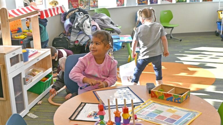 Young girl in pink shirt engaged in learning activity at classroom table with educational materials, other children playing in background of inclusive learning environment