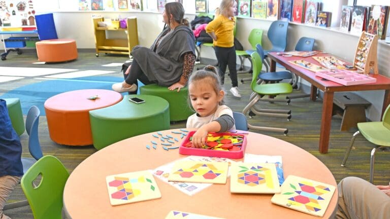 Young child engaged in shape-matching activity at colorful table in inclusive early intervention classroom with educators and other children present