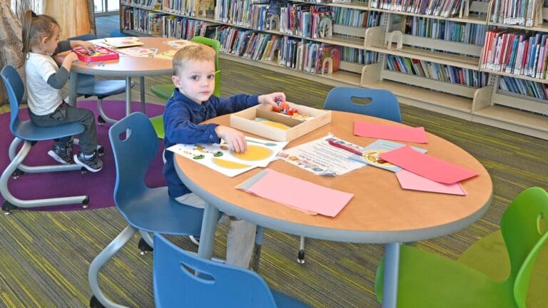 Young child in blue jacket engaged in creative activity at round table in library, with shelves of books visible in background