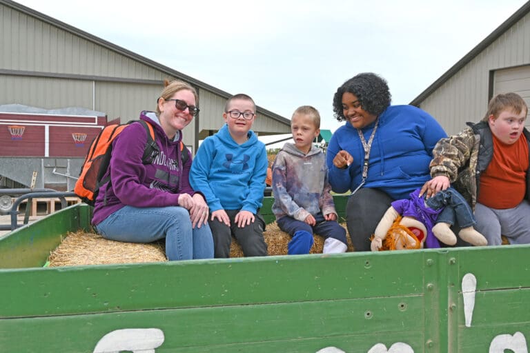 A woman and five children sit together in a green farm wagon filled with straw, smiling at the camera in front of red farm buildings in rural Ohio
