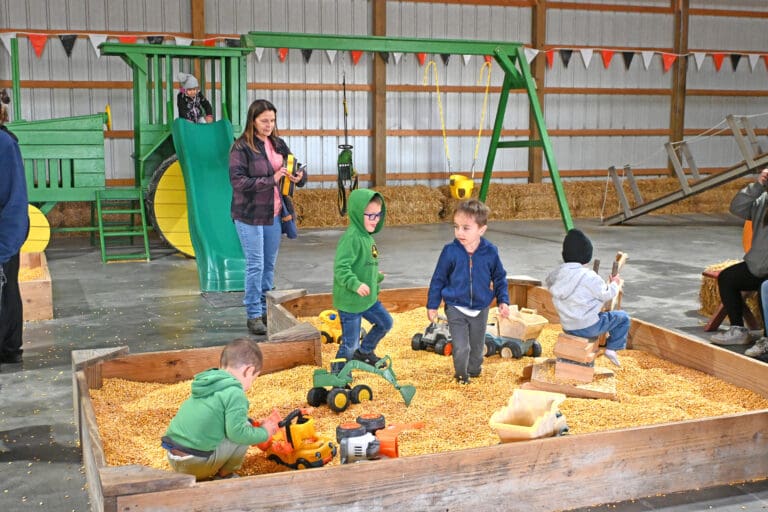 Children playing in a sandbox with toy construction vehicles at an indoor facility in Sandusky County, Ohio, with playground equipment and hay bales visible in the background