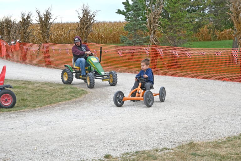 Child in orange go-kart and adult on green pedal tractor racing on dirt track with orange safety fencing at rural farm venue in Ohio