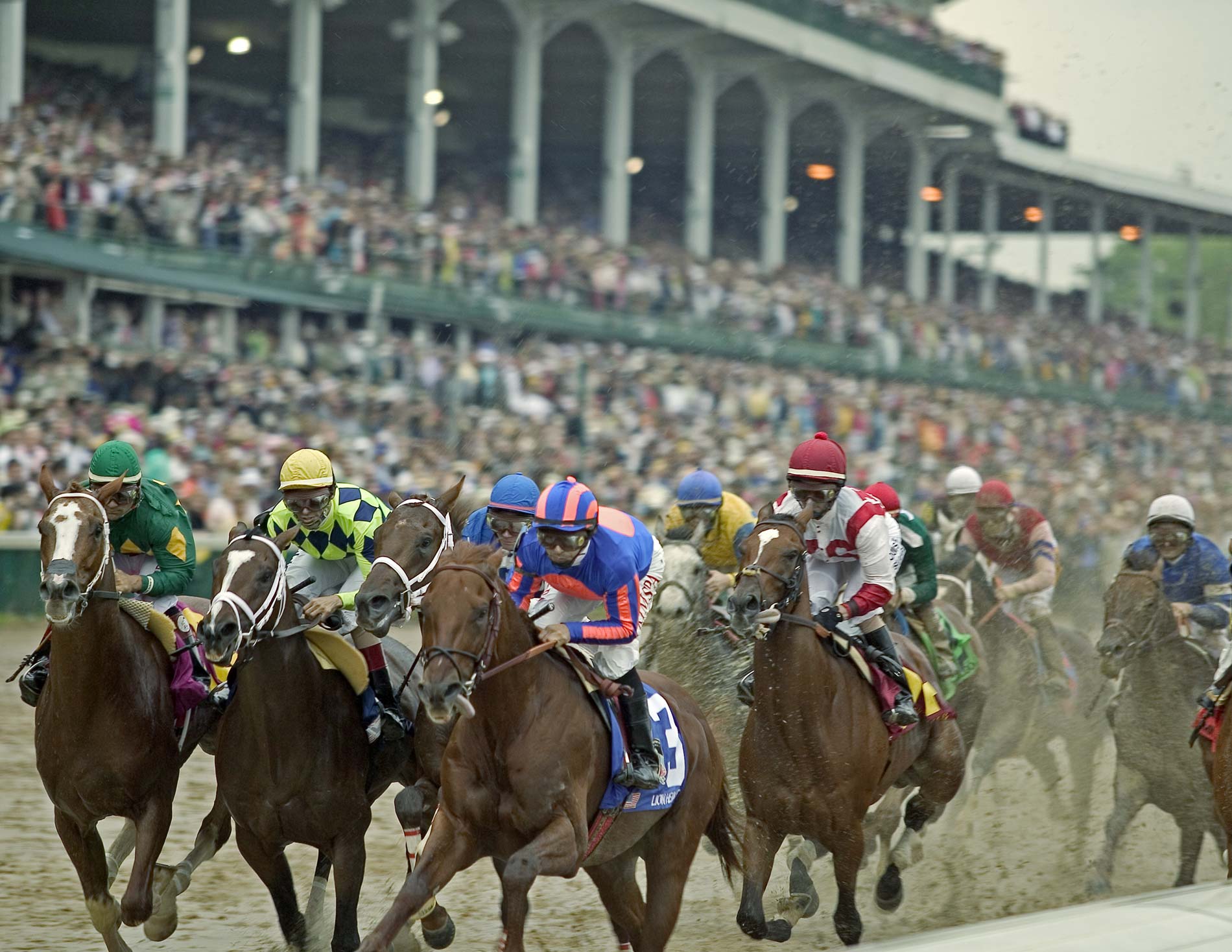 Thoroughbred racehorses with colorful jockeys compete on muddy track during Kentucky Derby with packed stadium grandstands in background