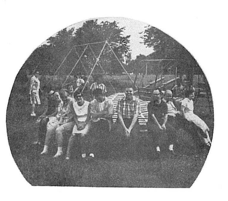 Black and white photograph of a group of people seated outdoors at a recreational facility with playground equipment and trees in the background, Sandusky County Ohio