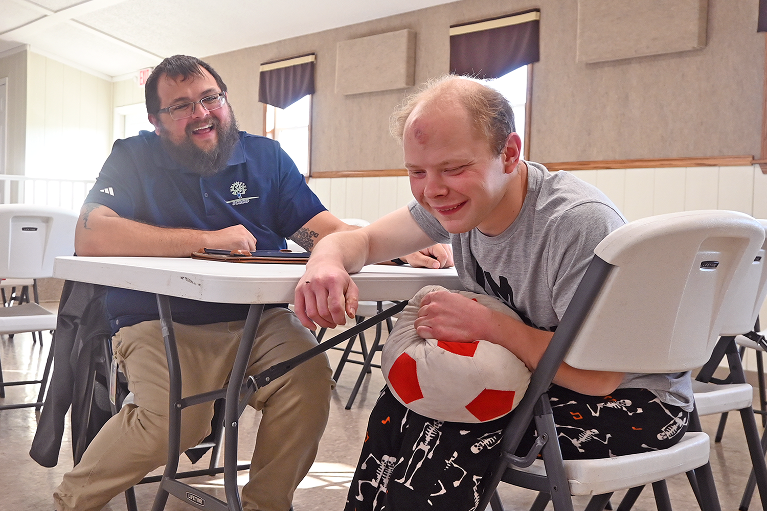 Man in blue polo shirt assists young man holding red and white soccer ball during developmental disability services activity in Sandusky County, Ohio