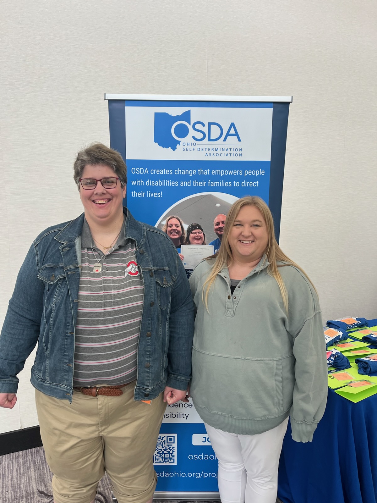 Two young adults smiling at OSDA event in Ohio, standing before banner for Ohio Self Determination Association promoting disability empowerment and family services